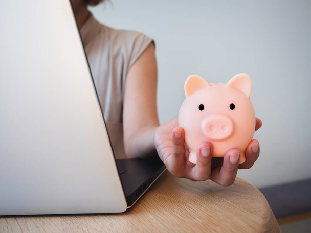 Money box on hand. Hand holding the pink piggy bank while using laptop computer on desk. Saving money wealth and financial concept, Business, online banking, finance, investment, financial planning. - Photo, Image