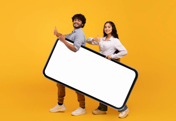 Happy indian couple holding huge smartphone with mockup for design, advertising app or website, showing thumbs up - Foto, afbeelding
