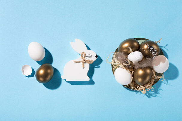 お祭りイースターの休日の挨拶カードの背景。イースターの白い黄金の卵、青いテーブルの上のウズラの卵と羽。スペースをコピーし、上のビュー。最小限のイースターカードのコンセプト、ハードシャドウ. - 写真・画像