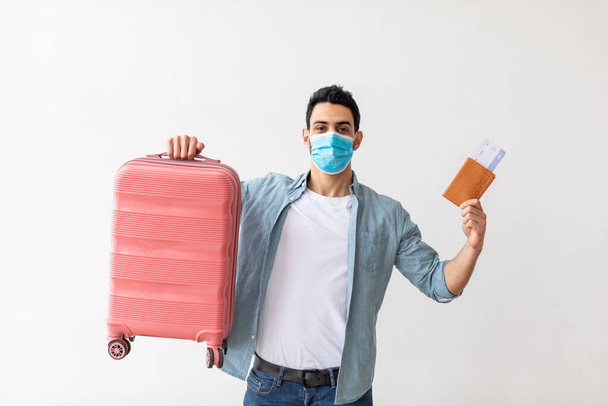 Arab man in medical mask holding suitcase and passport with airplane tickets, standing over light studio background - Foto, Bild