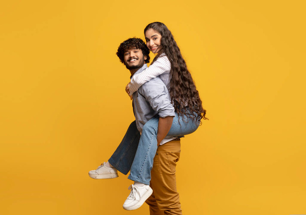 Young indian man giving piggyback ride to his girlfriend against yellow studio background, free space - Valokuva, kuva