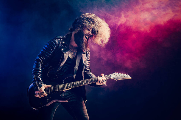 Emotional portrait of a rock guitar player with long hair and beard plays on the smoke background - Foto, imagen