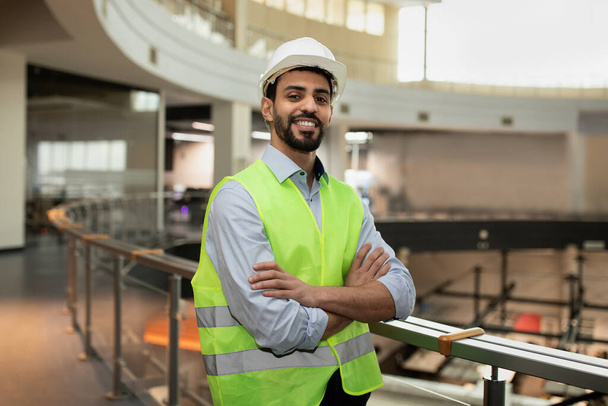 Gelukkige zelfverzekerde jonge aantrekkelijke Arabische ingenieur in harde hoed en beschermende kleding in kantoor plant interieur - Foto, afbeelding