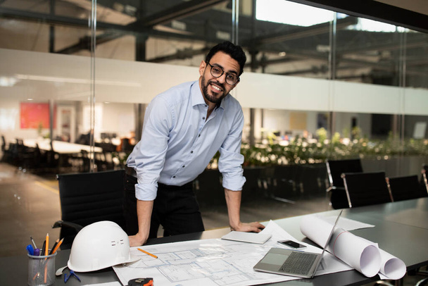Usmívající se tisíciletý atraktivní muslimský inženýr stavitel v brýlích pracující na stole s projektovými kresbami - Fotografie, Obrázek