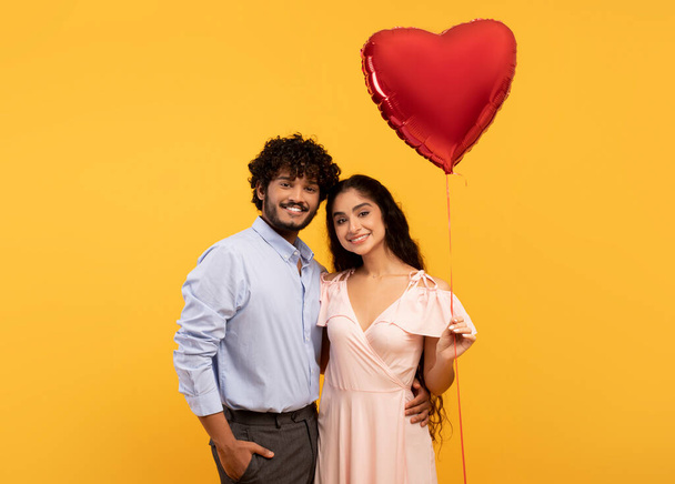 Loving indian couple holding big red heart shaped balloon, hugging and smiling at camera over yellow background - 写真・画像