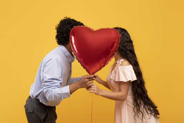 Romántica pareja india besándose detrás del globo en forma de corazón en el Día de San Valentín, posando sobre fondo amarillo, vista lateral - Foto, imagen
