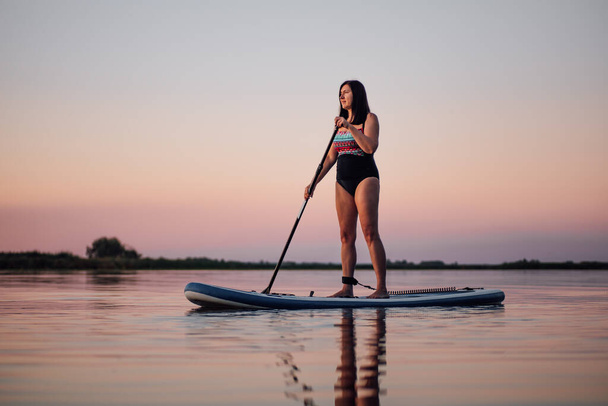 Relaxed middle aged woman rowing on sup board with oar looking at sunset on lake rippled water with pink sky in background. Active lifestyle for older people - Фото, зображення