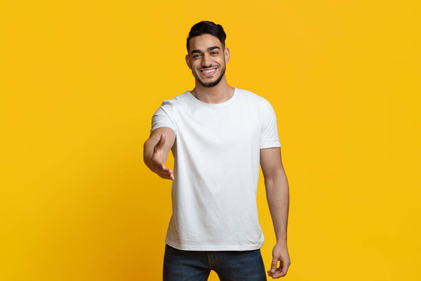 Charismatic young arabic guy outstretching hand on yellow - Foto, Imagem