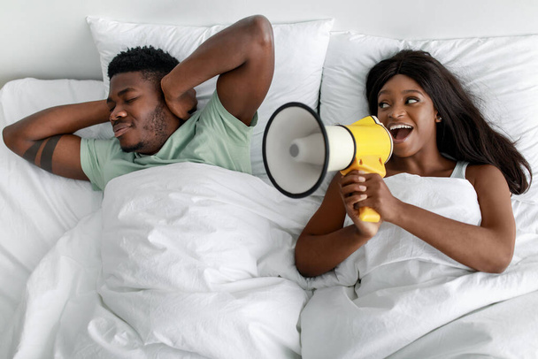 Smutny młody afrykański facet zamyka uszy na białym łóżku, wesoła kobieta krzyczy na megafon w sypialni - Zdjęcie, obraz