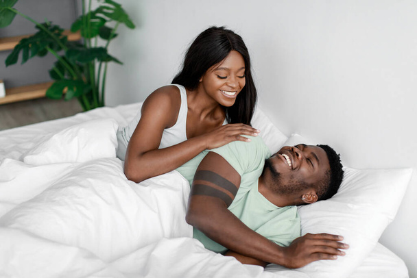 Smiling young pretty african american woman wakes up her husband after night sleep on bed in bedroom - Foto, afbeelding
