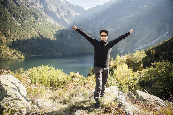 Elleri dağın tepesinde olan şapkalı genç turist doğaya hayran. - Fotoğraf, Görsel