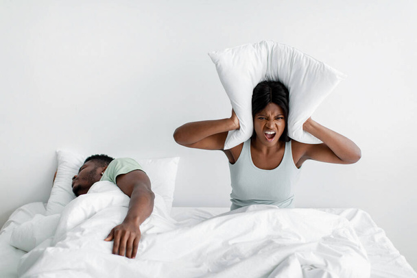 Unhappy young african american lady freaks out, screams, covers ears with pillow on bed, suffers from husband snoring - Foto, afbeelding