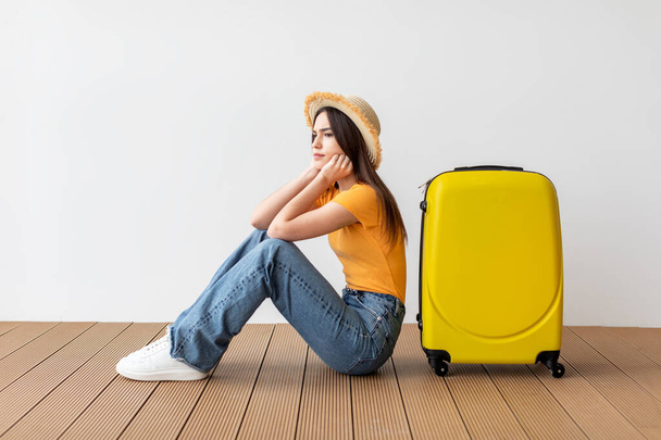 Aufgebrachte Touristin sitzt neben Koffer und blickt vor hellem Wandhintergrund in freien Raum - Foto, Bild