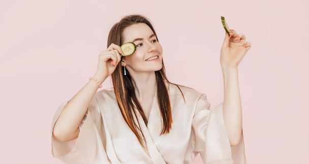 Girl make homemade face beauty masks. Cucumbers for the freshness of the skin around the eyes. Woman take care of youthful skin. Model laughing and having fun in spa on pink background - Foto, immagini
