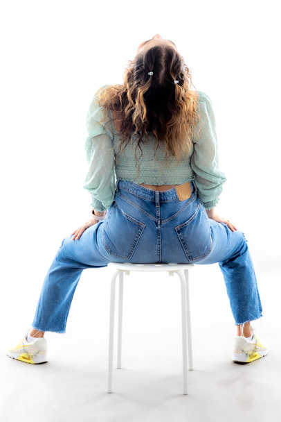 Latina teenage girl sitting on a small stool with her head tilted back, her back to the camera with a white studio background, long brown hair and casual clothes - Photo, image