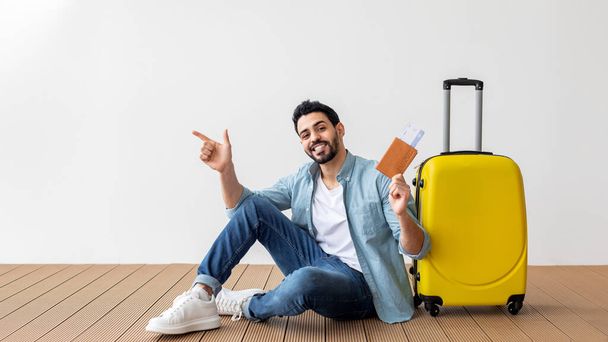 飛行機のチケットとパスポートを持っている幸せなアラブ人男性は、無料のスペースを指して、軽い壁の上にスーツケースの近くに座って - 写真・画像