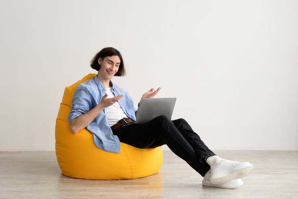 Externe communicatie. Jonge freelancer guy het maken van video call op laptop, zittend in zitzak stoel - Foto, afbeelding