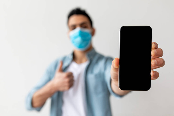 Arab guy showing smartphone blank screen and gesturing thumb up, wearing protective face mask, selective focus - Photo, image
