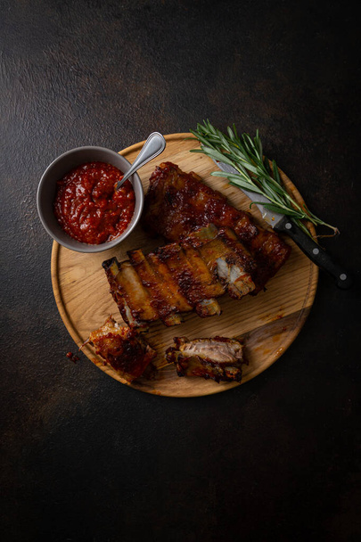Overhead view of grilled ribs and bbq sauce on wooden plate on dark surface - Fotografie, Obrázek