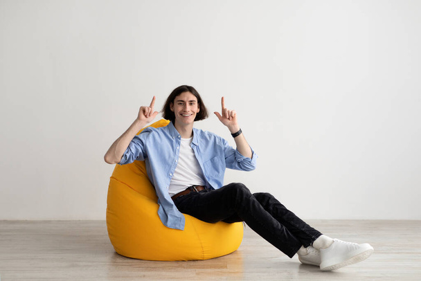Happy millennial cara apontando os dedos para cima e sorrindo para a câmera, publicidade oferta legal, sentado na cadeira beanbag - Foto, Imagem