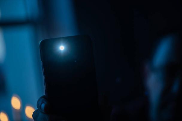 Mano masculina sostiene un teléfono inteligente con una linterna en llamas con ventana de iluminación en el fondo. - Foto, Imagen