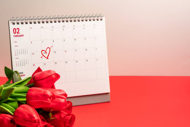 A koncepció Valentin nap február 14-én 2022. Jelölje meg a szív február 14-én naptár piros tulipán virág a piros háttér egy asztalon, Másolás tér, Szelektív fókusz, elmosódott háttér - Fotó, kép