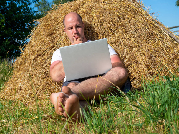 Tişört ve şort giymiş orta yaşlı bir adam elinde gri bir dizüstü bilgisayarla sarı bir samanlıkta yatıyor. Uzaktan çalışma, eğitim. Kırsal bölge. Düşünceli yüz ifadesi, duygu. - Fotoğraf, Görsel