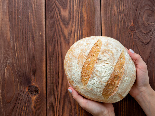 Крупним планом руки тримають круглий цілий бухан пшеничного хліба. Коричневий дерев'яний фон, вид зверху, плоский. Концепція хлібобулочних виробів. Копіювати простір
 - Фото, зображення