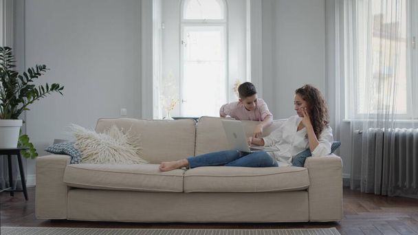 Young Mother And Son. Young Woman Lying On Sofa With Laptop. Beautiful Interior In Light Colors. - Photo, image