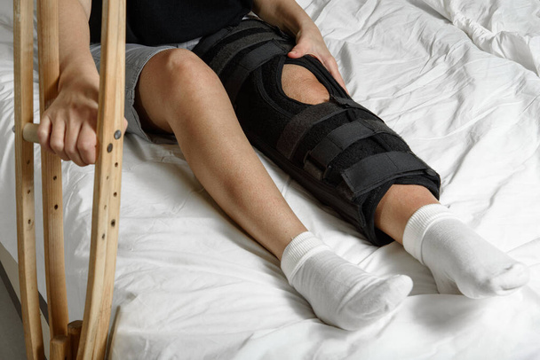 Patientin mit Orthese sitzt nach der Operation am gebrochenen Bein mit Krücken im Bett. Rehabilitation der verletzten Frau zu Hause. Selektiver Fokus - Foto, Bild
