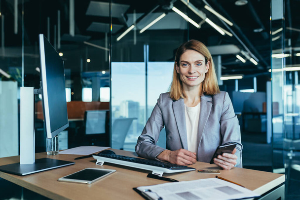 Portrait of experienced and successful business woman, boss manager holding phone looking at camera and smiling, woman working in modern office - Photo, image