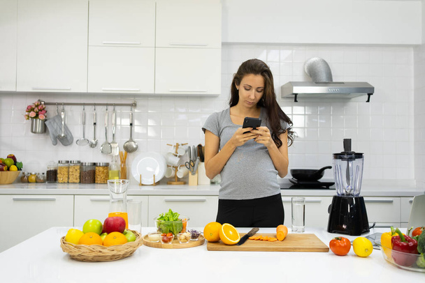 Schwangere trägt Kopfhörer und nutzt Smartphone in der Küche - Foto, Bild