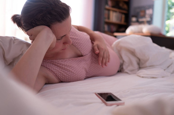 Biała kobieta z telefonem, która ma skurcze porodowe na kanapie w domu i oddycha - Zdjęcie, obraz