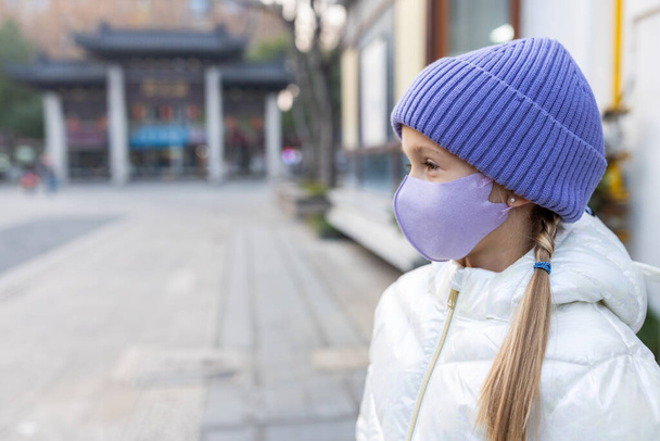 Cute little caucasian girl eight years old with blonde hair and protective face mask walking outdoor. Kid wearing stylish shirt and knitted hat violet color. Trendy color of the 2022 year very peri - Photo, Image