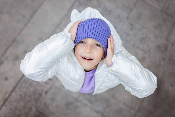 Cute little caucasian girl eight years old with blonde hair smiling outdoor. Kid wearing stylish shirt and knitted hat violet color. Trendy color of the 2022 year very peri - Fotoğraf, Görsel