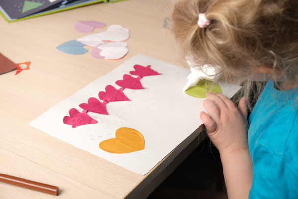 preschooler girl in blue t-shirt makes applique sitting at the table, hearts cut out of colored bougue for card for Valentine's Day, children's creativity, atopic dermatitis on the hands of a child - Photo, image