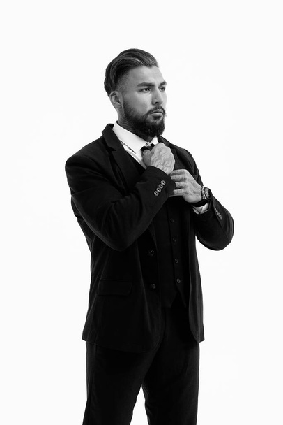 Hombre barbudo hispano seguro con las manos tatuadas en camisa blanca y pantalones negros ajustando la corbata y mirando hacia otro lado mientras está de pie sobre fondo gris - Foto, imagen