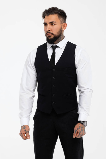 Chico hispano barbudo con chaleco oscuro y camisa blanca con corbata mirando a la cámara con las manos en los bolsillos en el estudio - Foto, Imagen