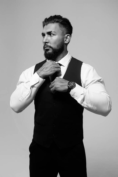 Confident Hispanic bearded man with tattooed hands in white shirt and black trousers adjusting tie and looking away while standing against gray background - Fotó, kép