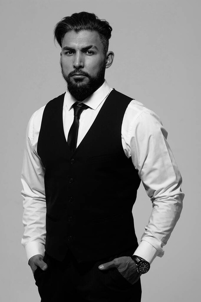 Bearded Hispanic guy in dark vest and white shirt with tie looking at camera with hands in pockets in studio - Фото, зображення
