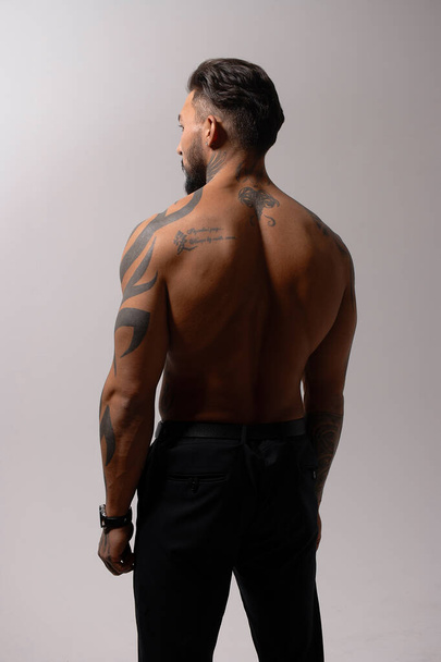 Hispanic shirtless male model with muscular tattooed torso standing with hands in pockets and looking away on gray backdrop - 写真・画像