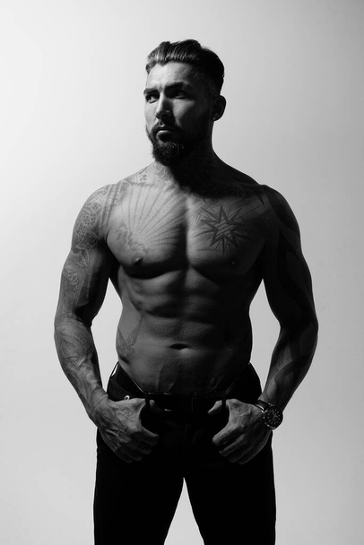 Hispanic shirtless male model with muscular tattooed torso standing with hands in pockets and looking away on gray backdrop - Fotografie, Obrázek