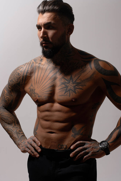 Hispanic shirtless male model with muscular tattooed torso standing with hands in pockets and looking away on gray backdrop - Fotó, kép