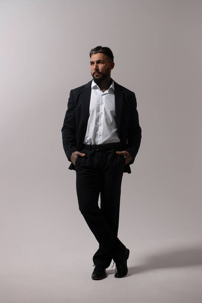 Bearded Hispanic guy in dark vest and white shirt with tie looking at camera with hands in pockets in studio - Foto, imagen