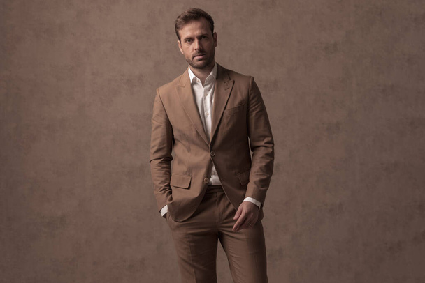 attractive businessman holding one hand in pocket and posing against brown background - Fotoğraf, Görsel