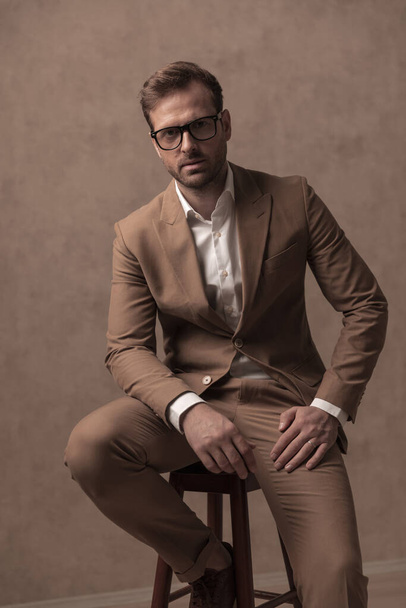 sexy macho businessman resting on a chair and wearing eyeglasses in a fashion pose - Fotoğraf, Görsel