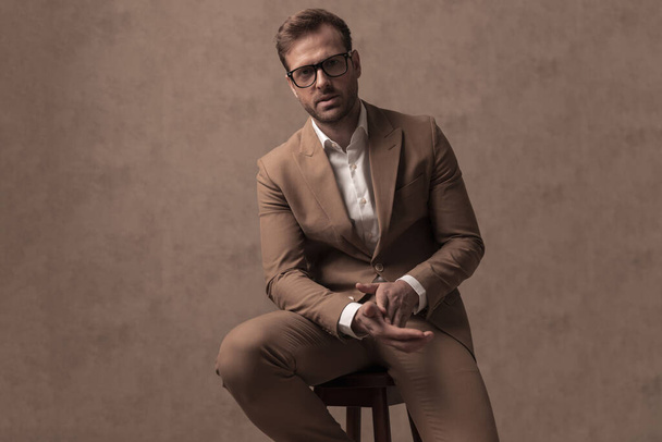 attractive fashion businessman rubbing his palms, wearing eyeglasses and sitting against brown background - Zdjęcie, obraz
