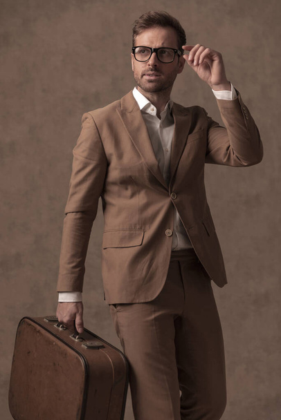 sexy hombre de negocios de moda posando mientras fija sus gafas y con un maletín en la mano  - Foto, Imagen