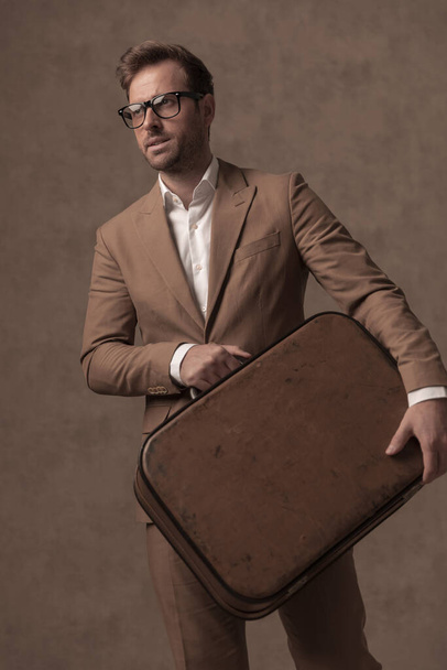 sexy businessman posing with his briefcase in hands, wearing eyeglasses and a suit in a fashion pose - Fotografie, Obrázek