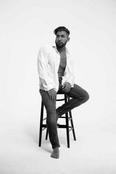 Modello maschile ispanico senza maglietta con busto muscoloso tatuato in piedi con le mani in tasca e distogliendo lo sguardo sullo sfondo grigio - Foto, immagini
